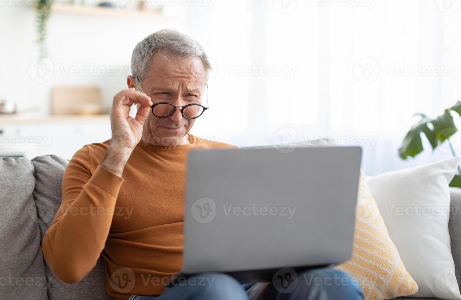 Poor Eyesight. Senior Man Squinting Eyes Using Laptop Wearing Eyeglasses Having Problems With Vision Sitting On Couch. Ophtalmic Issue, Bad Sight In Older Age, Macular Degeneration Concept photo