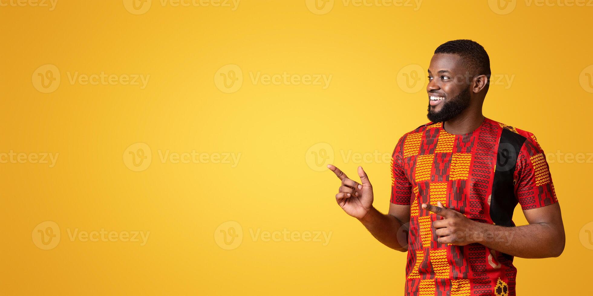 Excited black guy in african costume pointing at free space for advertisement, yellow background, panorama photo