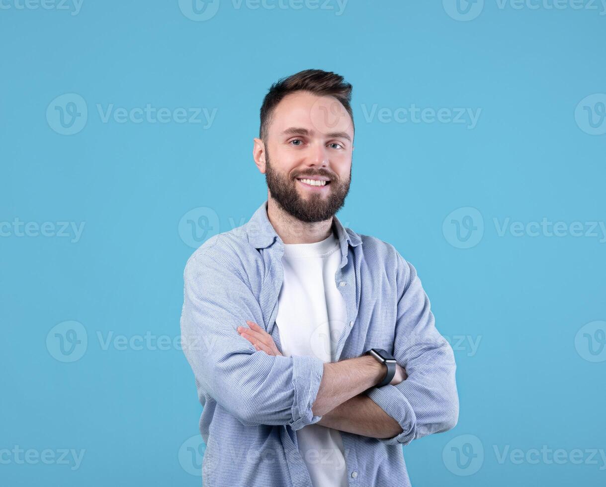 Portrait of positive young guy posing with crossed arms and smiling at camera on blue studio background. Masculine bearded millennial man in casual clothes having happy face expression photo