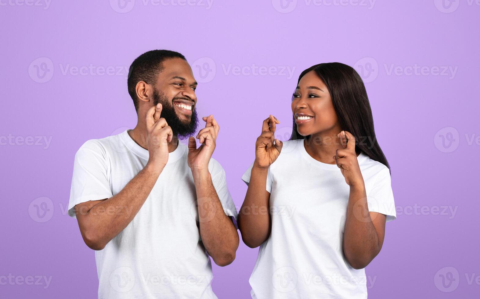 Making Cherished Wish. Positive Black Couple Crossing Fingers And Smiling, Begging For Luck, Praying For Miracle, Hoping For Good Things To Happen, Looking At Each Other Over Blue Studio Wall photo