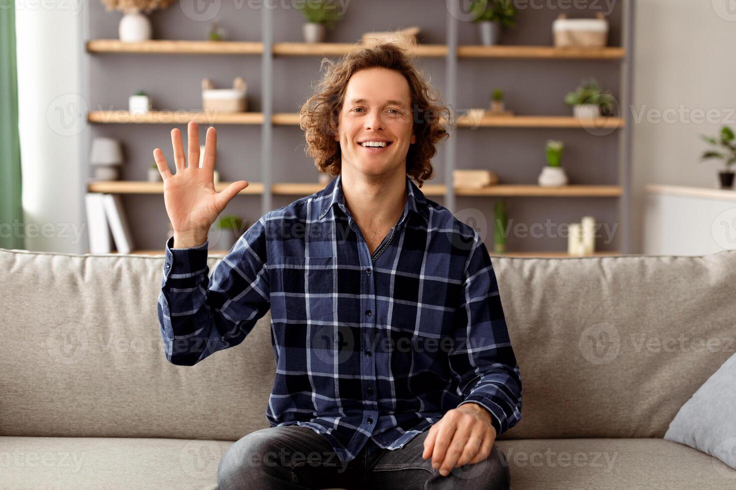 Hello Greeting. Cheerful Guy Waving Hand To Camera Sitting On Sofa At Home. Young Man Smiling And Gesturing Hi Posing Indoors. Call, Modern Distance Communication Concept photo