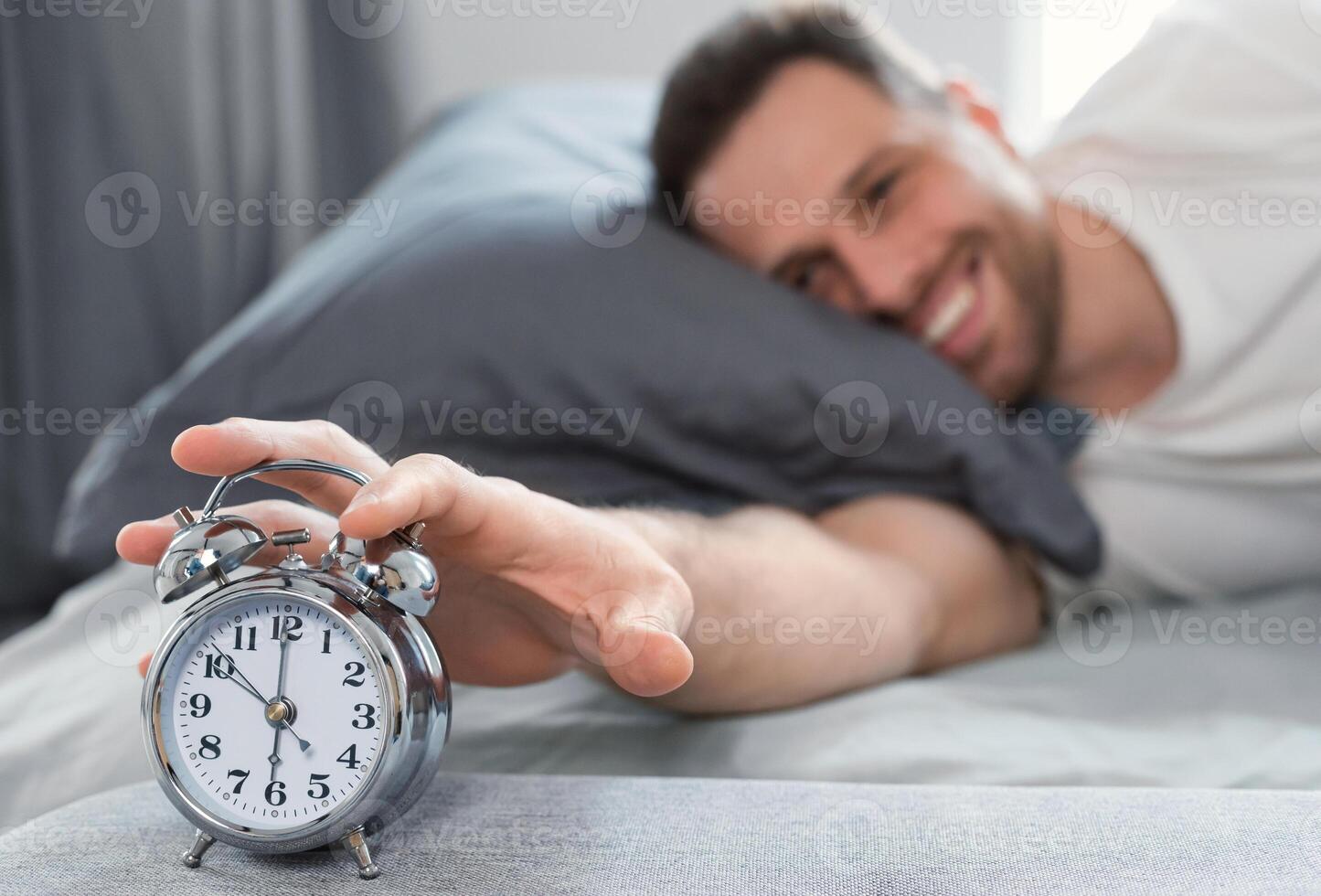 Good Morning. Happy Well-Slept Man Turning Off Alarm-Clock Waking Getting Up Lying In Bed At Home. Shallow Depth, Selective Focus photo