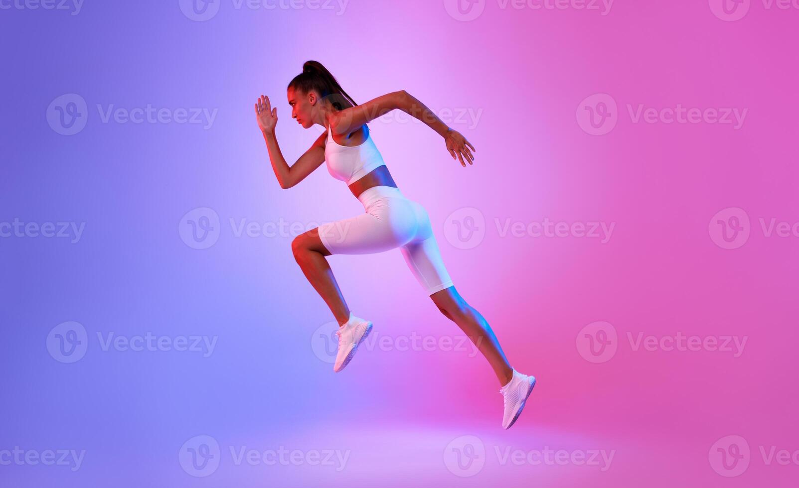 Determined Sporty Woman Running In Mid-Air Exercising During Cardio Workout Over Pink And Blue Neon Studio Background, Wearing White Fitwear. Fitness And Sport Concept. Side View, Full Length photo