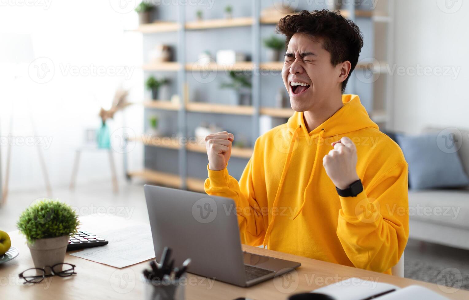 Excited asian man emotionally celebrating success using laptop in home office, sitting at desk raising hands with joy, shaking fists, received good news, got promotion, finished project, free space photo