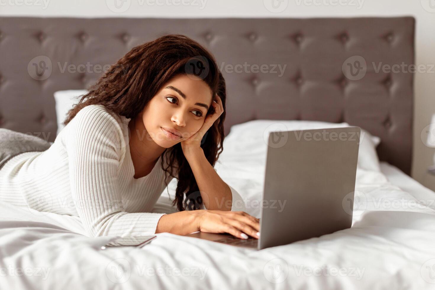 Portrait of African American woman lying in bed and using laptop computer, chatting or browsing social media. Young lady watching or movie, working remotely at home office, typing on keyboard photo