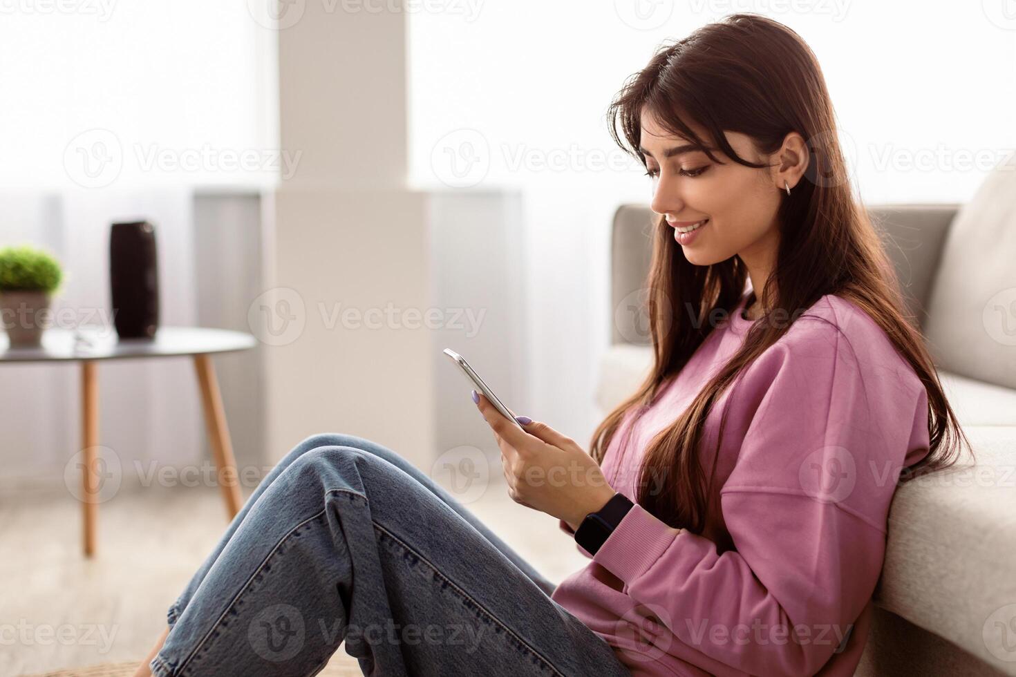 Cool Gadget And Application. Portrait of young smiling beautiful woman holding mobile phone, typing sms message, sitting and leaning on the sofa in living room. Woman browsing web, free space photo