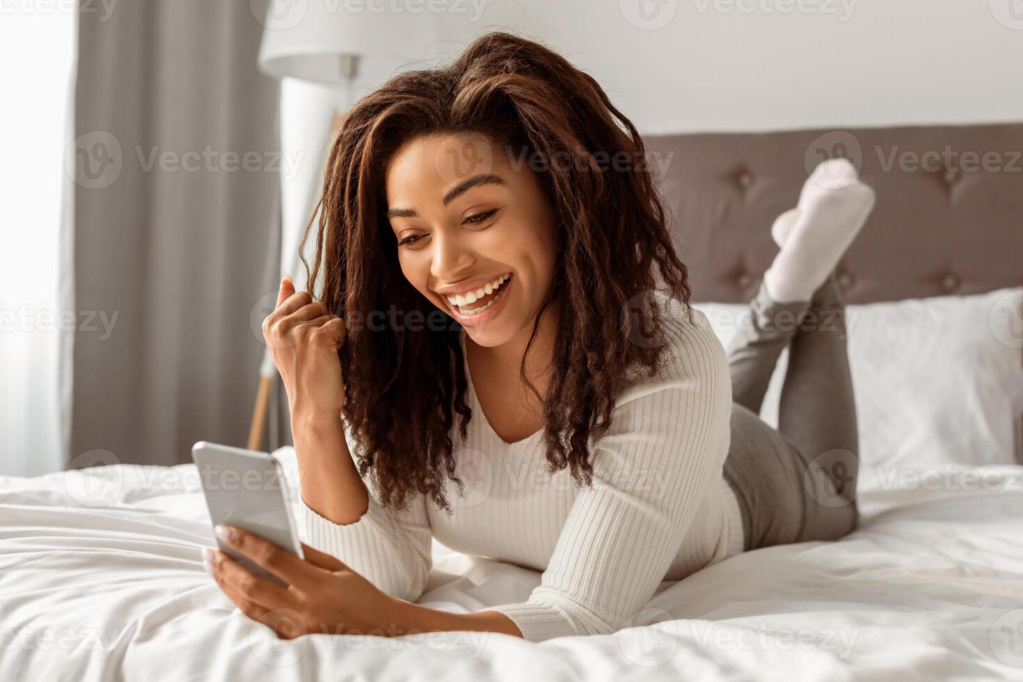 Big Win. Portrait Of Joyful African American Woman Holding Smartphone Shaking Fist In Joy And Excitement. Happy Young Female Lying In Bed At Home. Yeah, Great News And Message Concept photo
