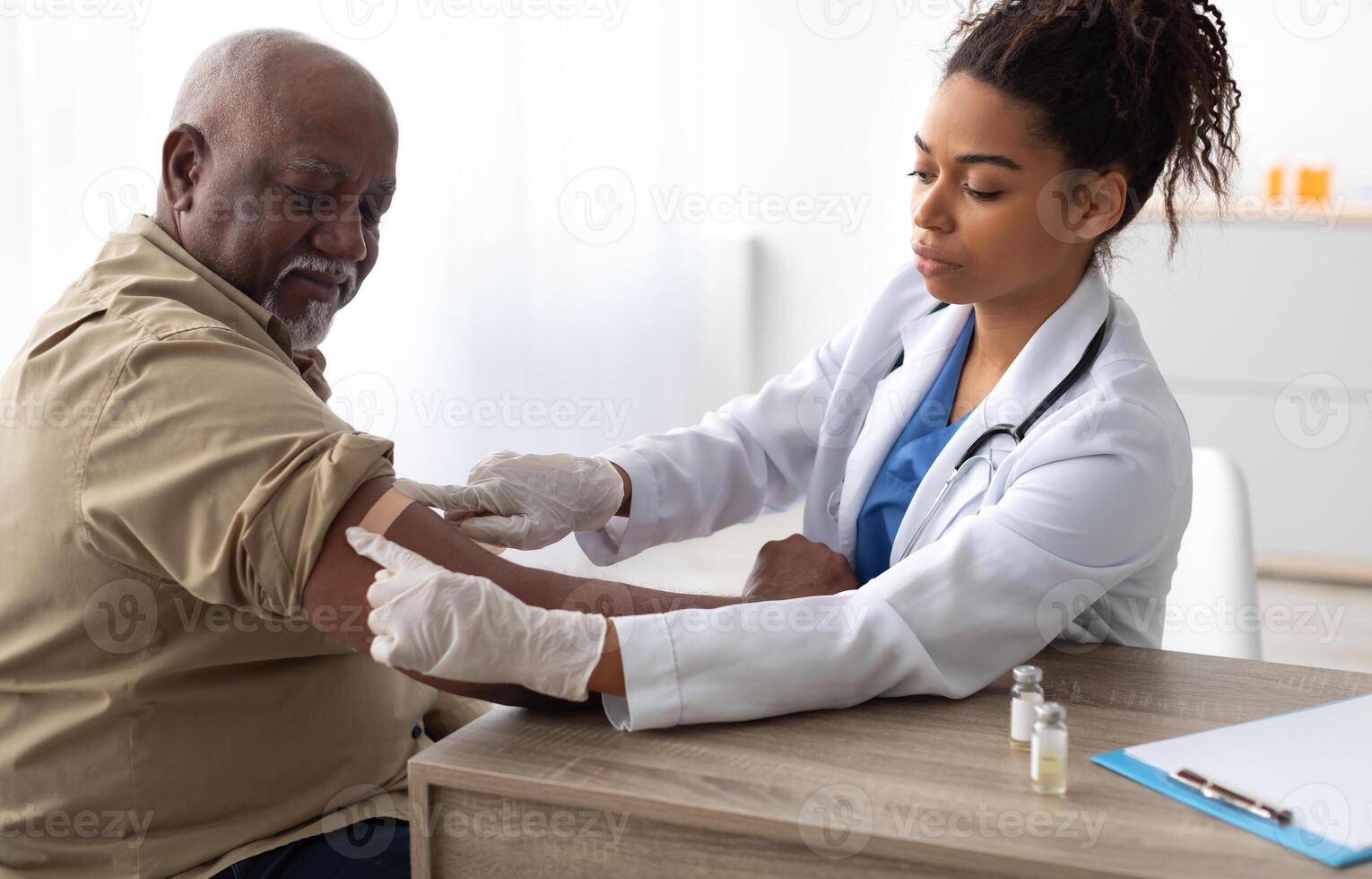 Vaccination Concept. Senior Black Man Getting Vaccinated Against Flu Or Corona Virus With Antiviral Vaccine, Female Doctor Putting Applying Sticking Adhesive Bandage On Arm After Injection In Hospital photo