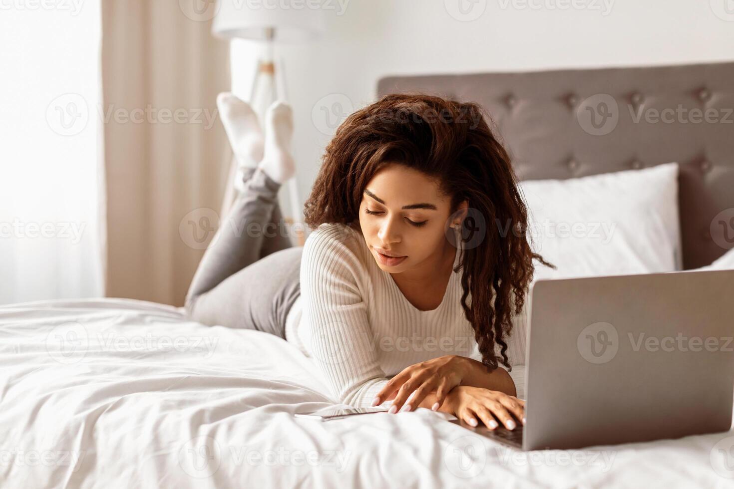 Always In Touch. Portrait of young African American woman lying in bed, using mobile phone and working on laptop computer. Curly black lady reading text message, free copy space photo