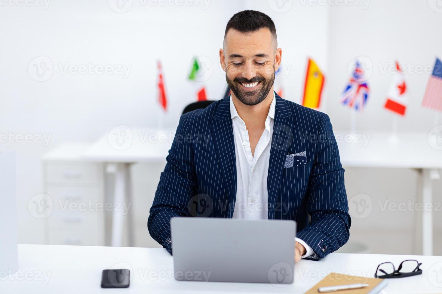 A confident man in a pinstripe suit smiles while typing on his laptop in a bright office. International flags decorate the background, emphasizing a global business environment. photo