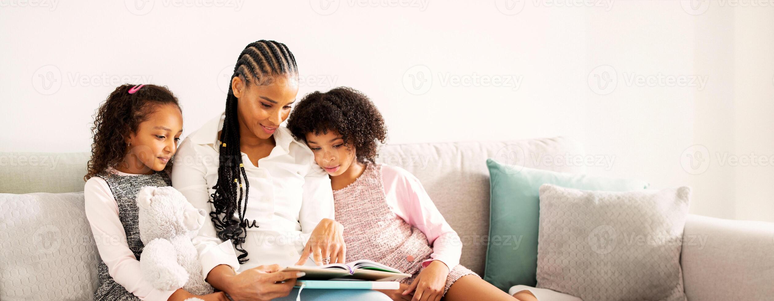 A black mother engages her two daughters with a story, sitting on a comfortable couch in a bright, inviting living room. The children show enthusiasm as they listen. photo