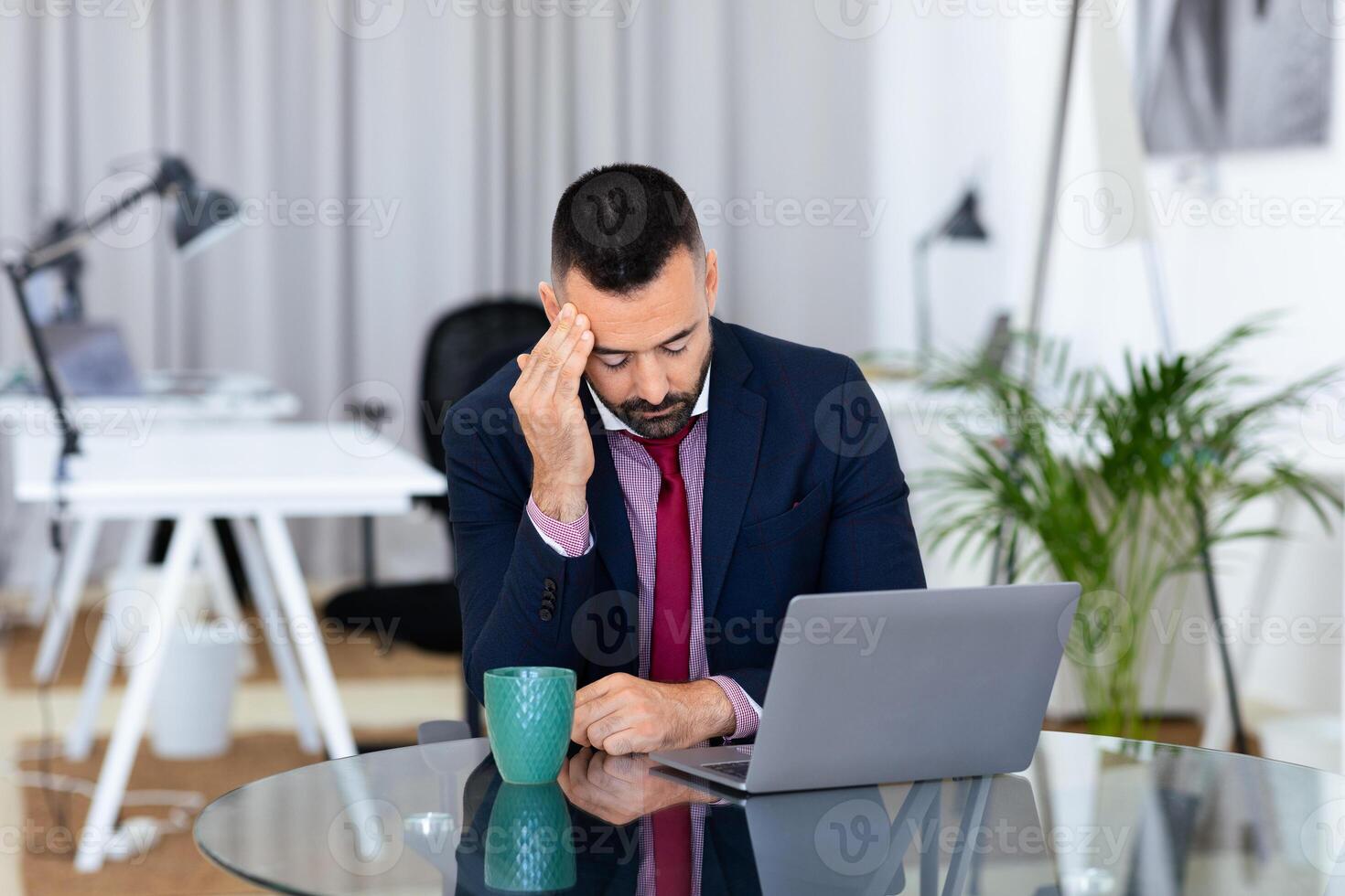 In a modern office, a businessman sits at a glass table, looking stressed while resting his forehead on his hand. A laptop is in front of him, and he holds a coffee mug in his other hand. photo