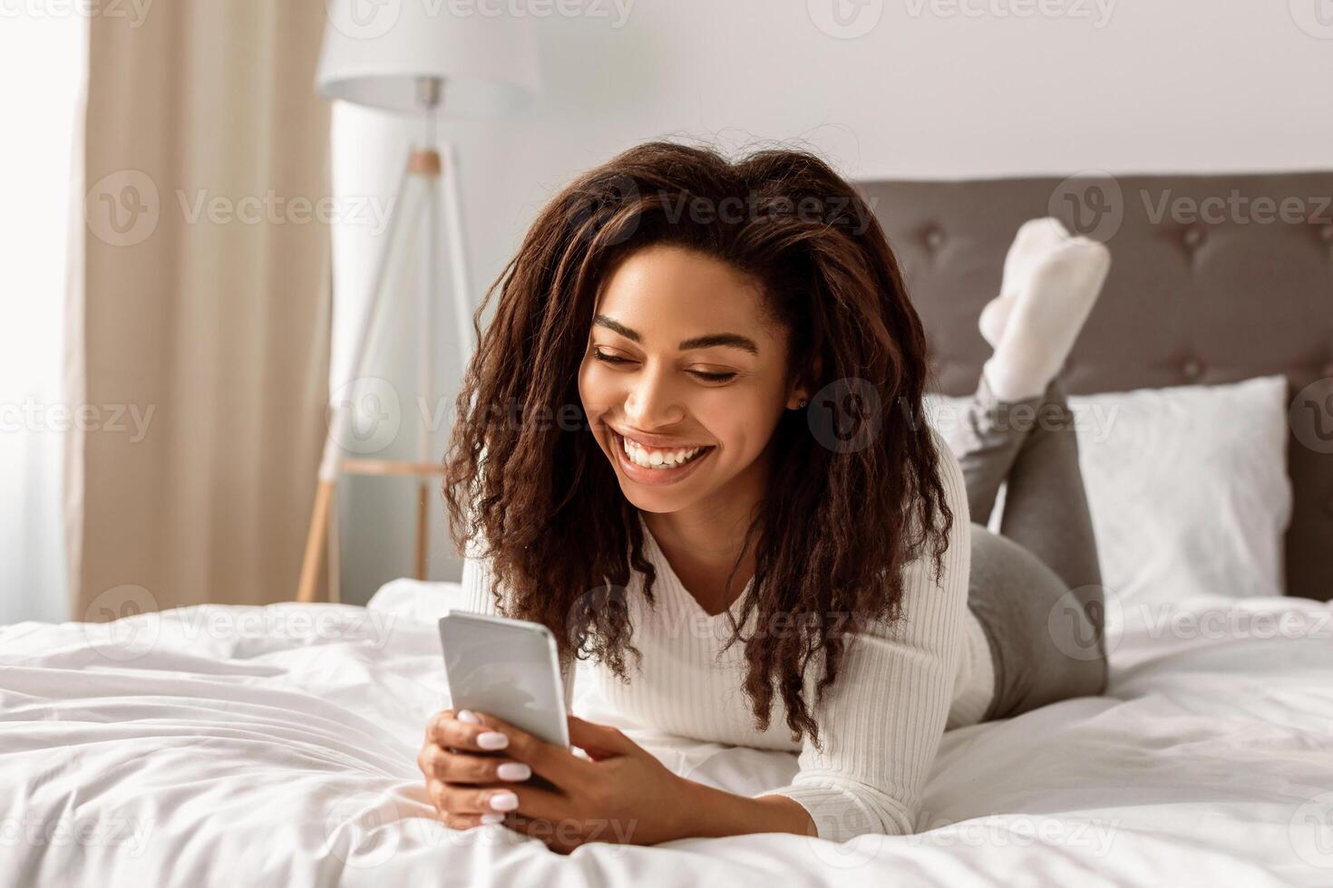 Mobile Phone User. Portrait of happy African American woman lying in bed and smiling while reading text message from someone, holding and using smartphone, chatting with lover or browsing social media photo