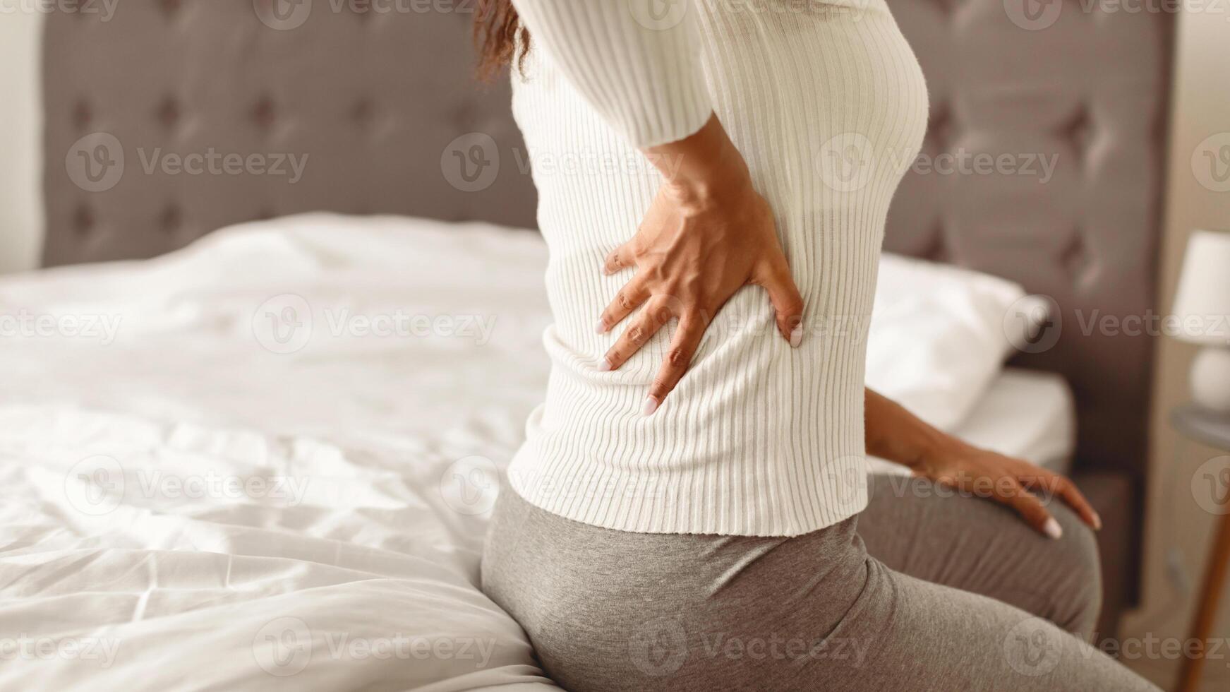 People and Health Problem Concept. Coseup of sick young black woman suffering from acute pain in left side, sitting on bed at home, touching lower back with hand, free copy space photo