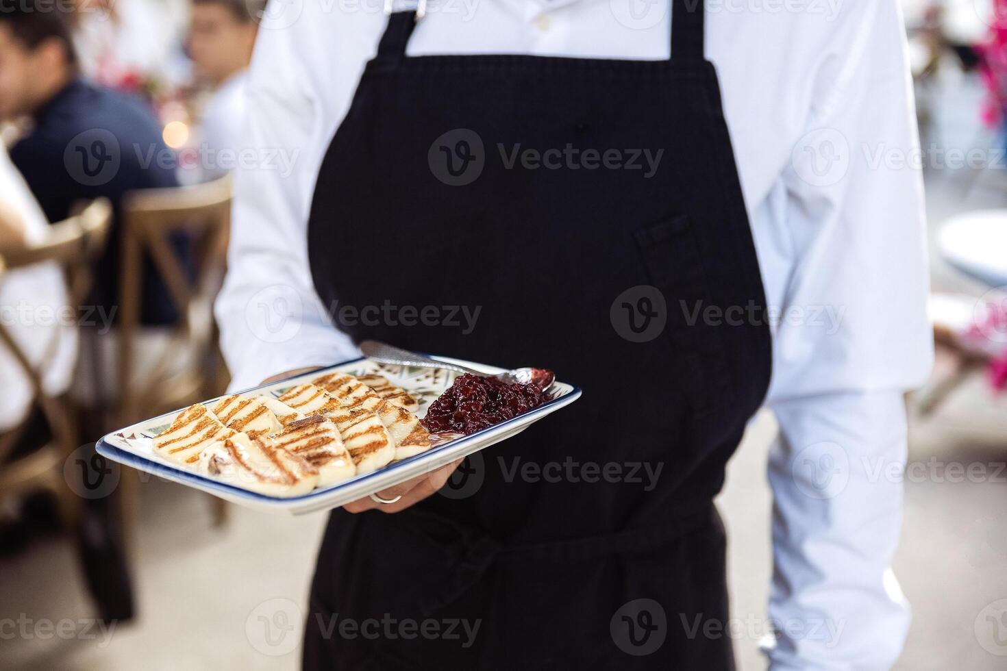 Close-up of waiter hands holding tray with fried soft cheese and jam. Man in black apron carries plate with traditional Cypriot grilled pickled cheese haloumi. photo