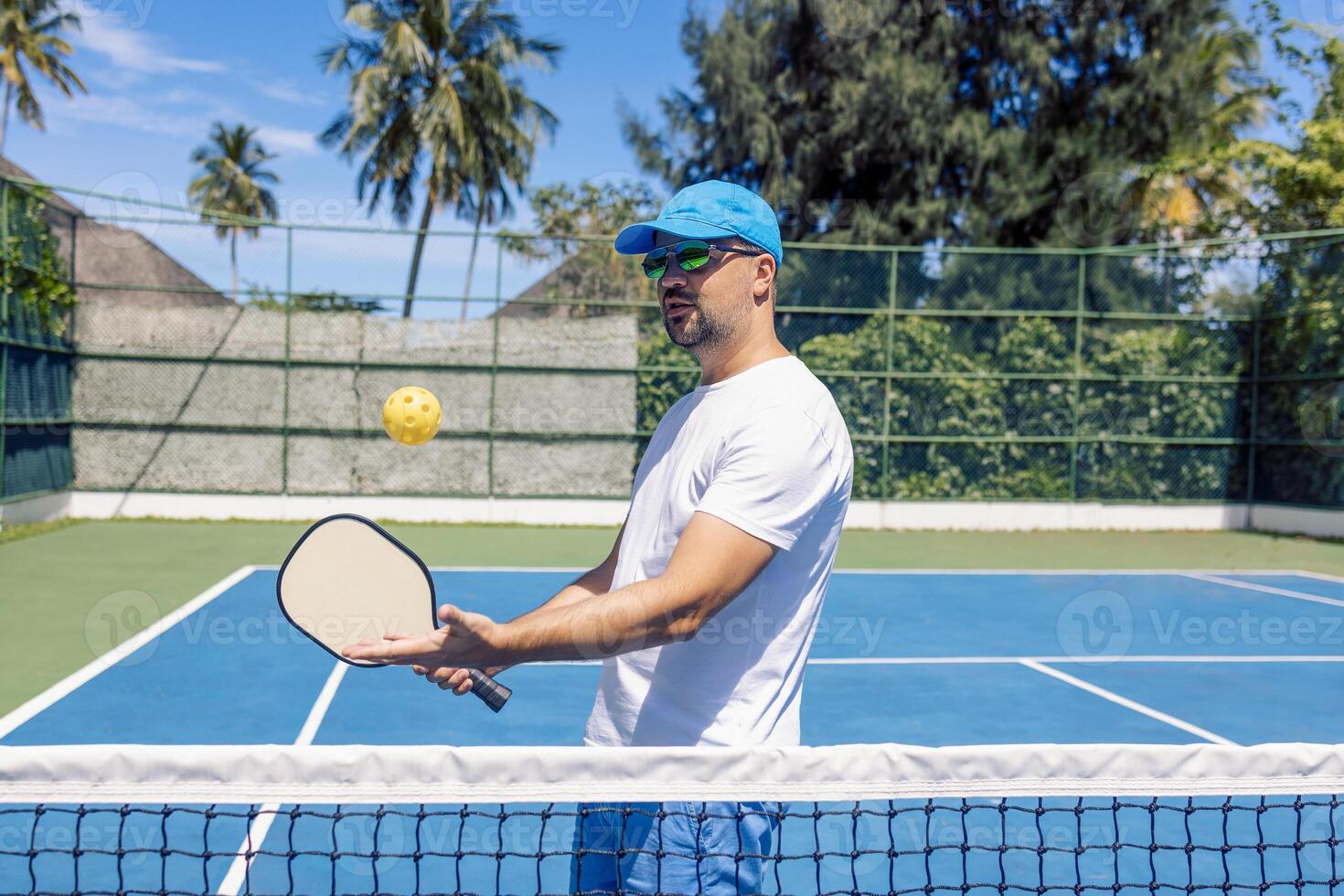 A young man in a cap and sunglasses throws a yellow ball and holds a racket. An athlete plays pickleball outdoors. photo