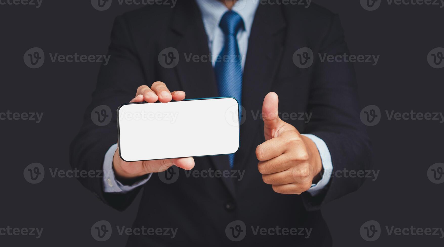 A businessman holding a horizontal mobile phone with a blank white screen and showing thumbs up while standing on a gray background photo