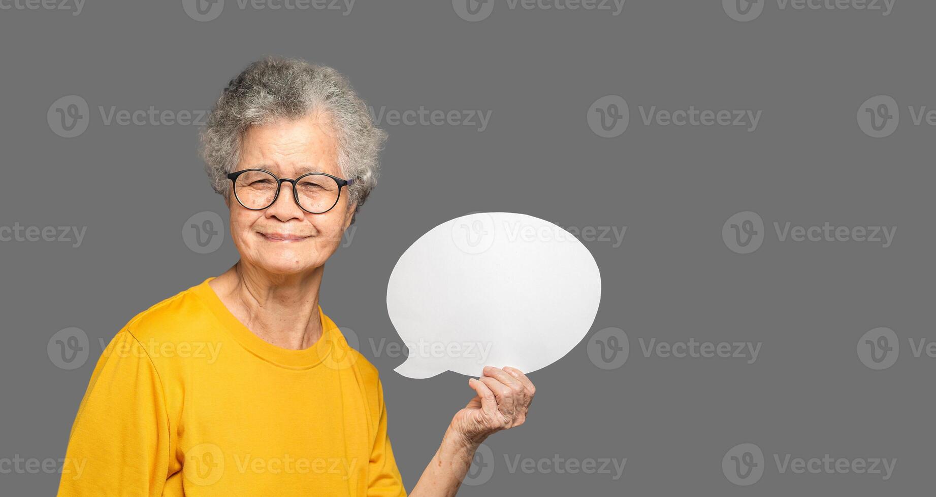 Senior woman in an orange casual holding a blank speech bubble and looking at the camera with a smile while standing on a gray background photo