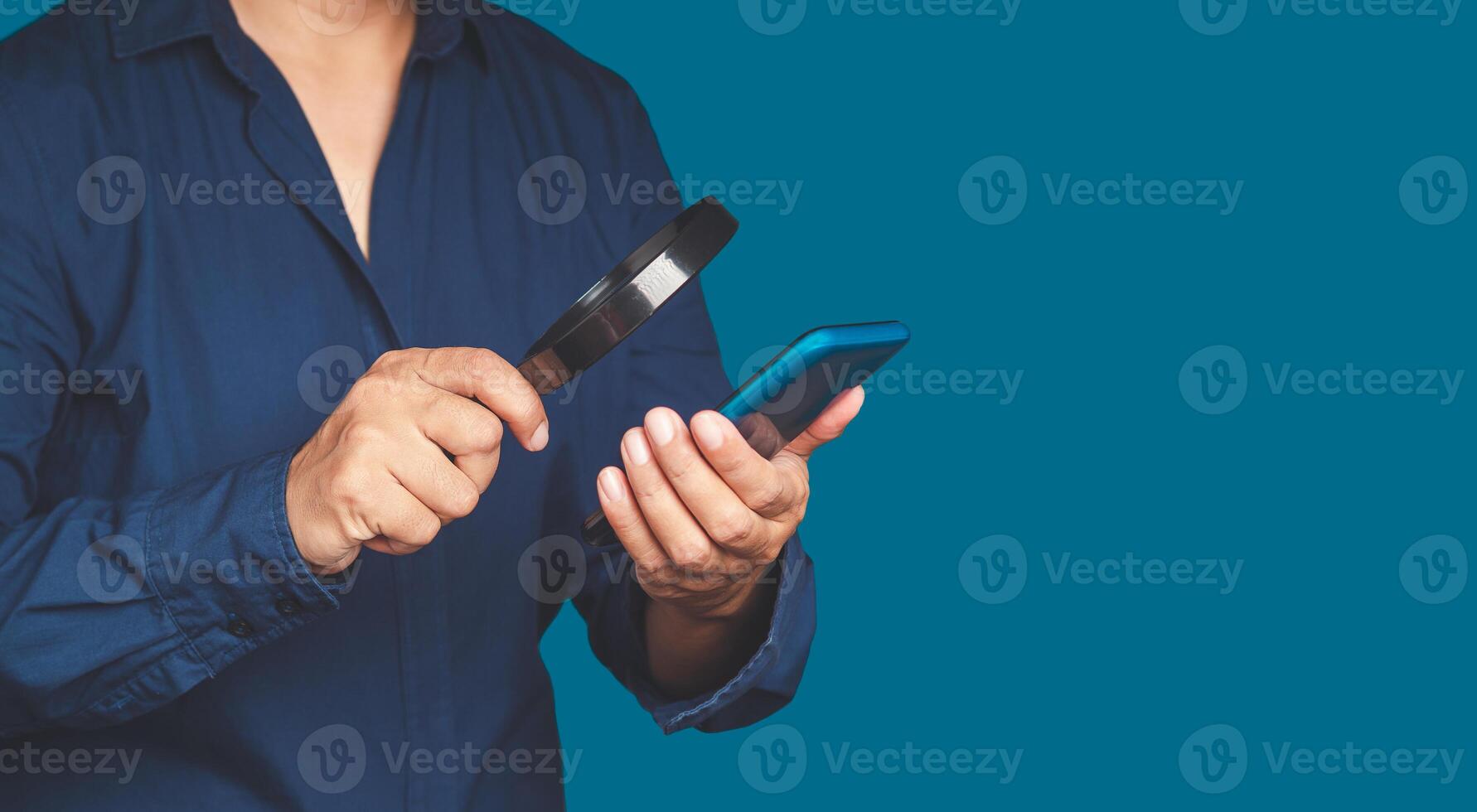 A businessman is holding a magnifying glass and looking at a smartphone while standing on a blue background photo