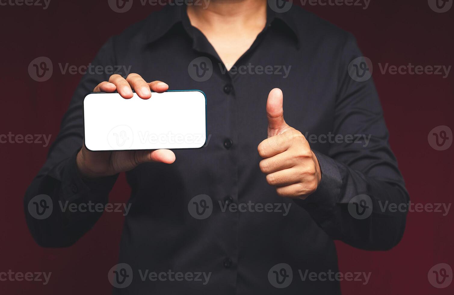 A businessman holding a horizontal mobile phone with a blank white screen and showing thumbs up while standing on a red background photo
