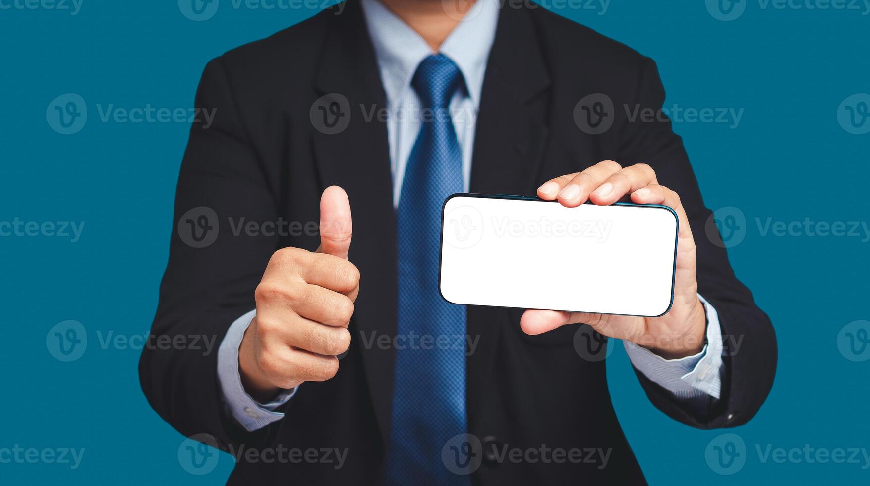 A businessman holding a horizontal mobile phone with a blank white screen and showing thumbs up while standing on a blue background photo