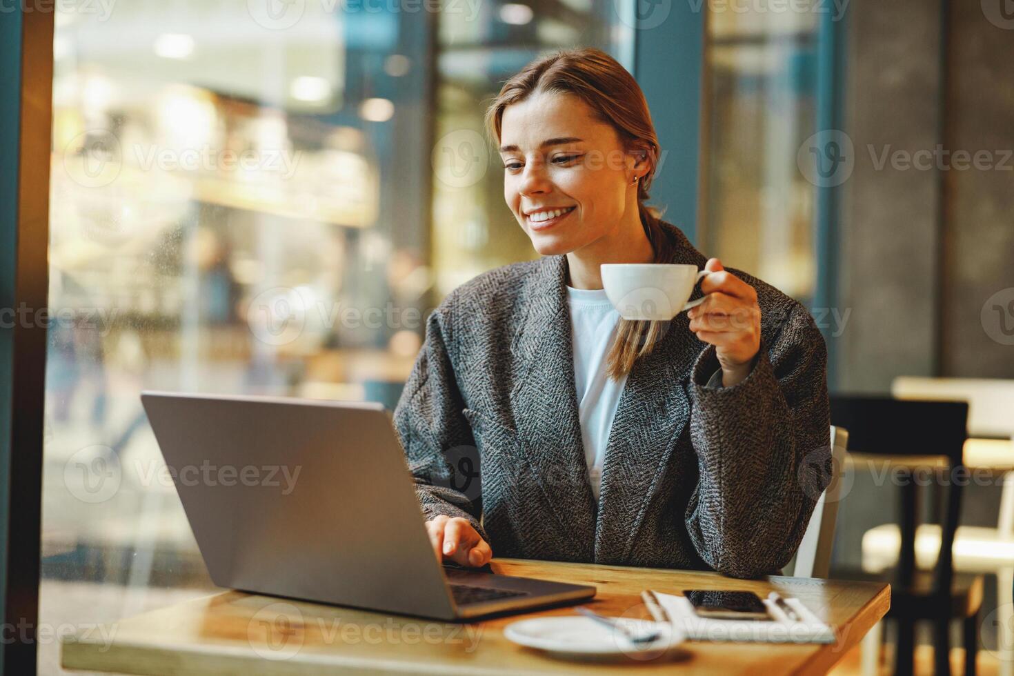 Smiling female entrepreneur is drinking coffee in coworking while working on laptop near window photo