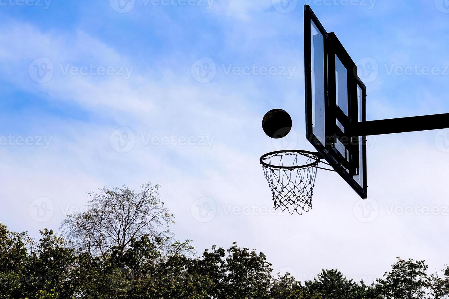 Silhouette of Basketball Hoop and Ball Against Blue Sky with Trees in the Background on a Clear Day photo