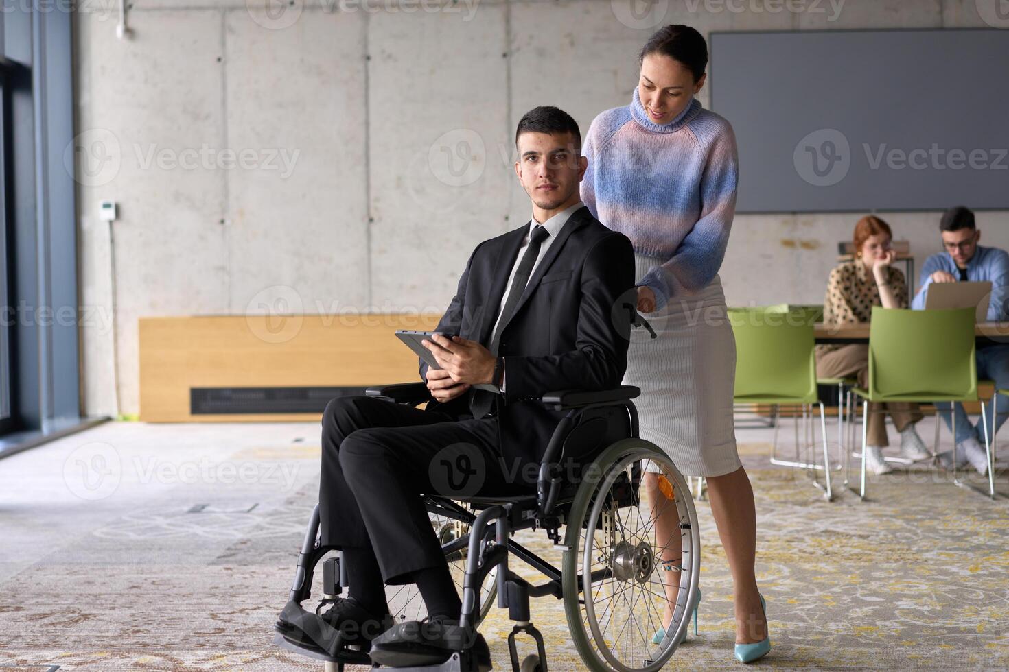 In a professional office setting, a businesswoman helping her director, who is sitting in a wheelchair and using a tablet, while their colleagues collaborate in the background. photo