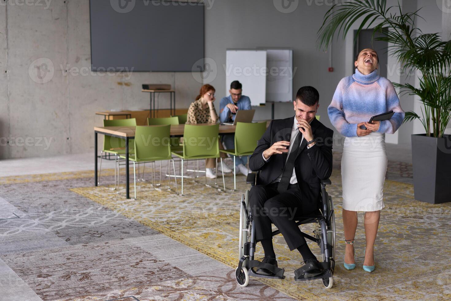In a professional office setting, a businesswoman helping her director, who is sitting in a wheelchair and using a tablet, while their colleagues collaborate in the background. photo
