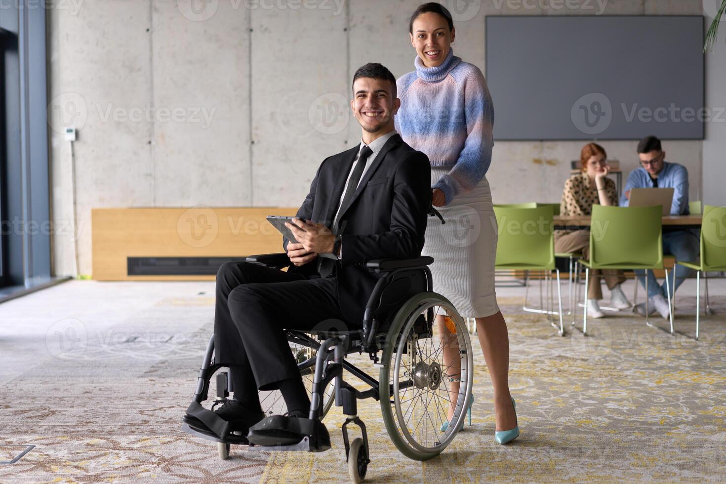 In a professional office setting, a businesswoman helping her director, who is sitting in a wheelchair and using a tablet, while their colleagues collaborate in the background. photo