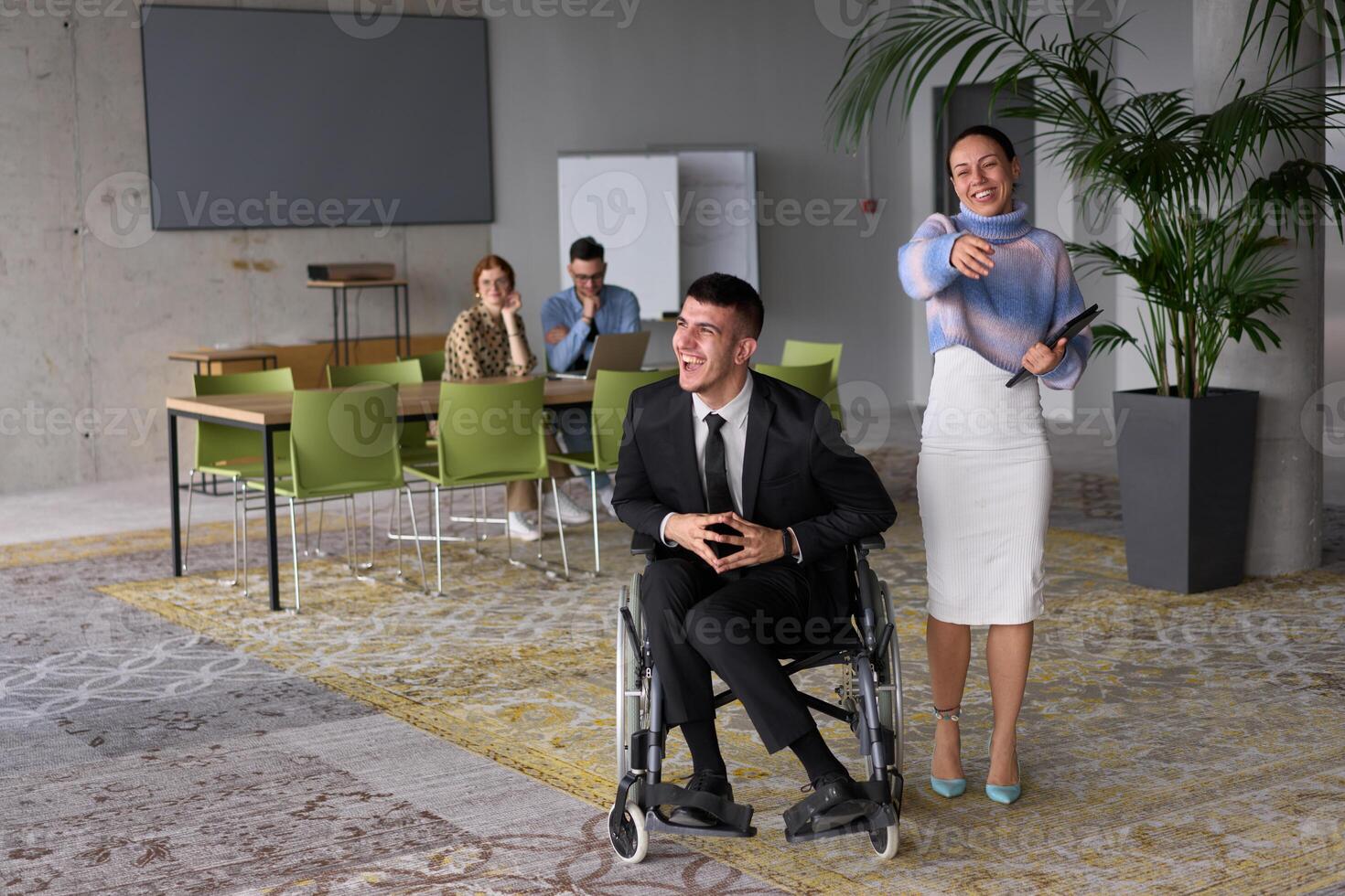 In a professional office setting, a businesswoman helping her director, who is sitting in a wheelchair and using a tablet, while their colleagues collaborate in the background. photo