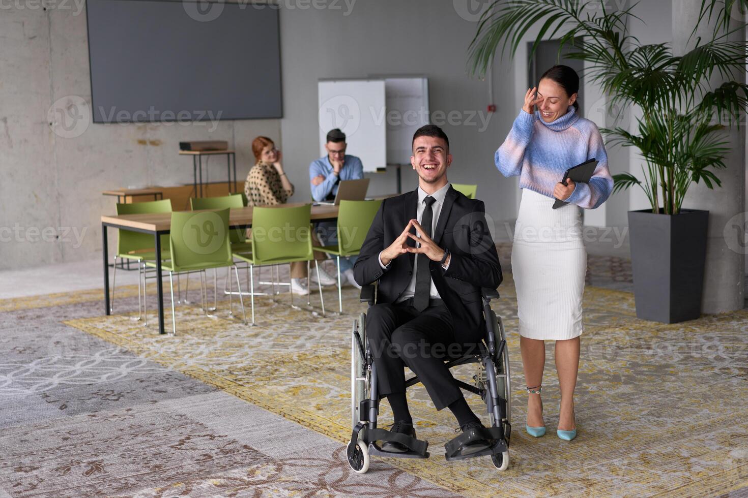 In a professional office setting, a businesswoman helping her director, who is sitting in a wheelchair and using a tablet, while their colleagues collaborate in the background. photo