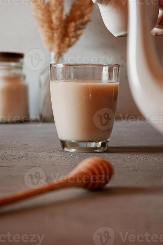 Herbal or black tea with milk in a glass cup. Serving with sugar, honey spoon and teapot on the table. Morning and breakfast hot beverage concept. Vertical photo