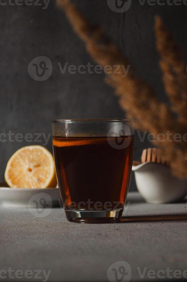 Glass with black tea and slice of lemon on the table, honey spoon and lemon in the background. Vertical photo. Breakfast and morning hot beverage concept. Dark mood photo