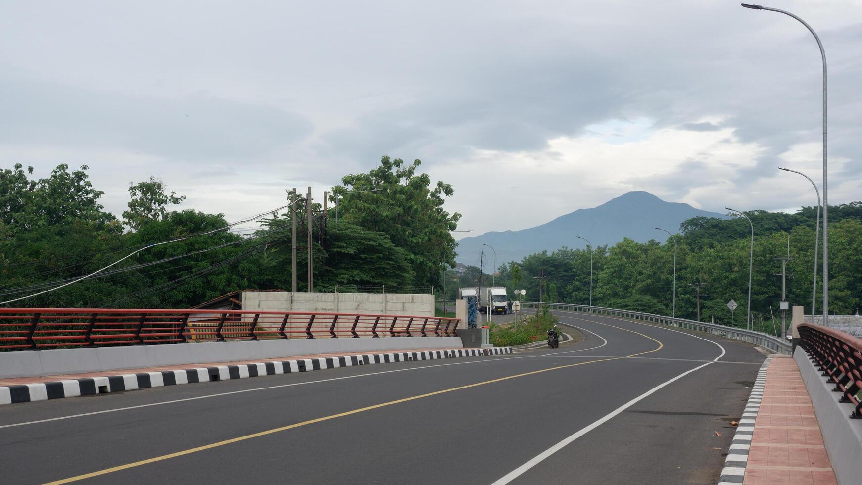Scenic Highway with Mountain View and Cloudy Sky, Surrounded by Green Trees and Lamp Posts photo