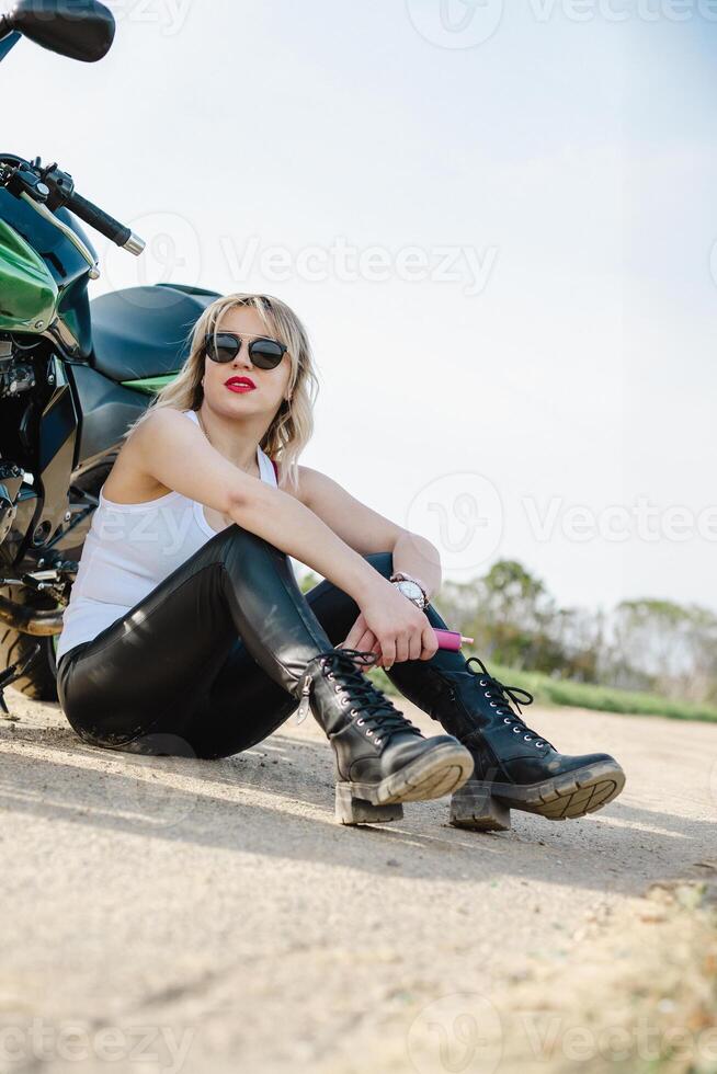 A woman in black pants and a white tank top sits casually on the ground beside a motorcycle. The image exudes a relaxed, adventurous vibe perfect for lifestyle and travel themes photo