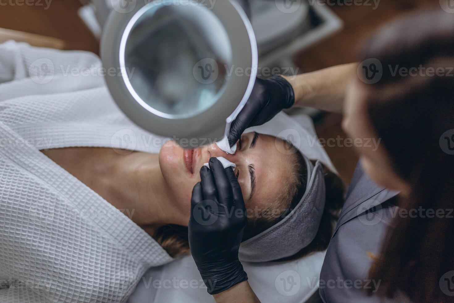 Cosmetologist in rubber gloves performs facial cleansing using a magnifying glass with LED light lupa photo