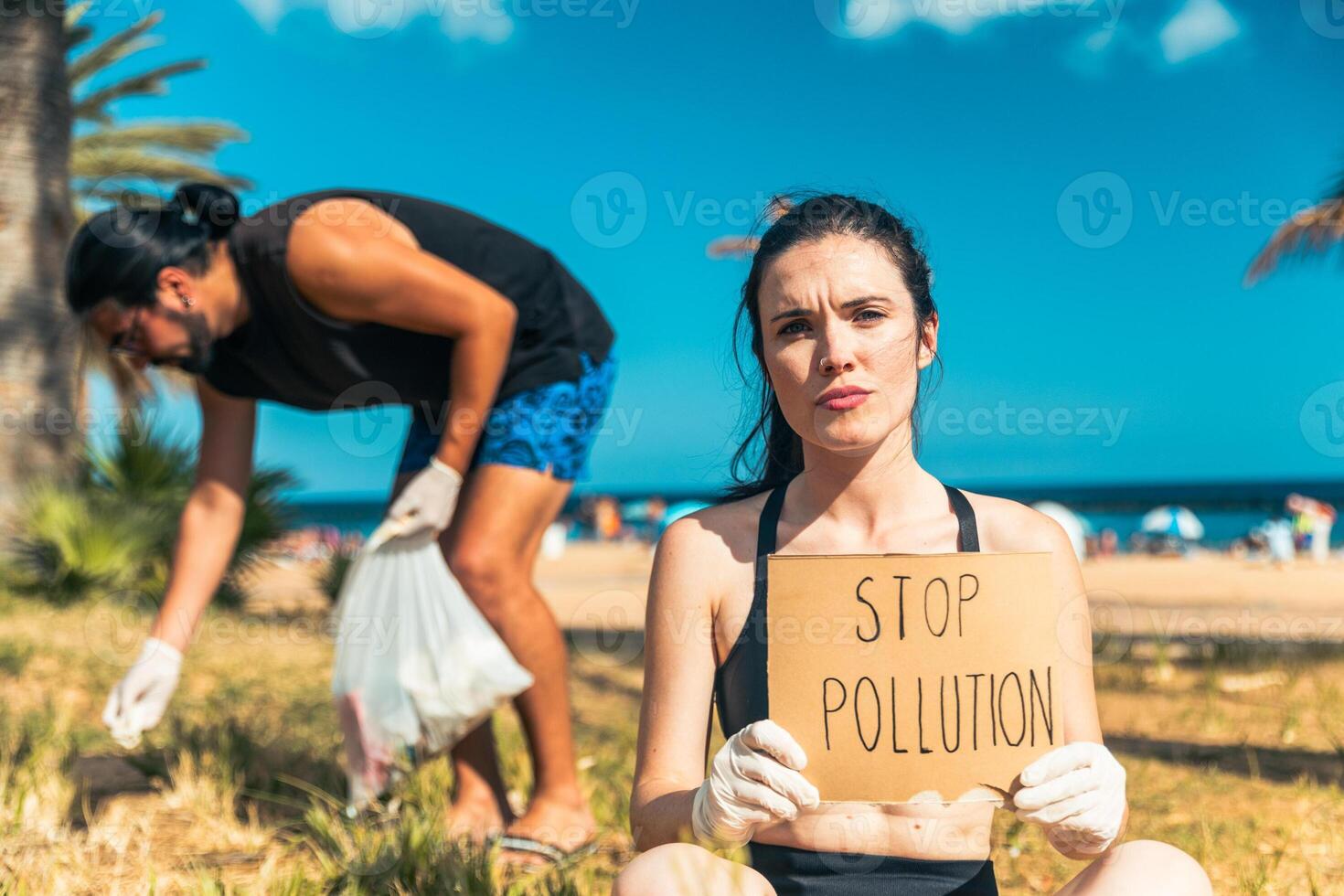 An activist is holding a sign that says Stop Pollution at the beach, her colleague collects trash photo