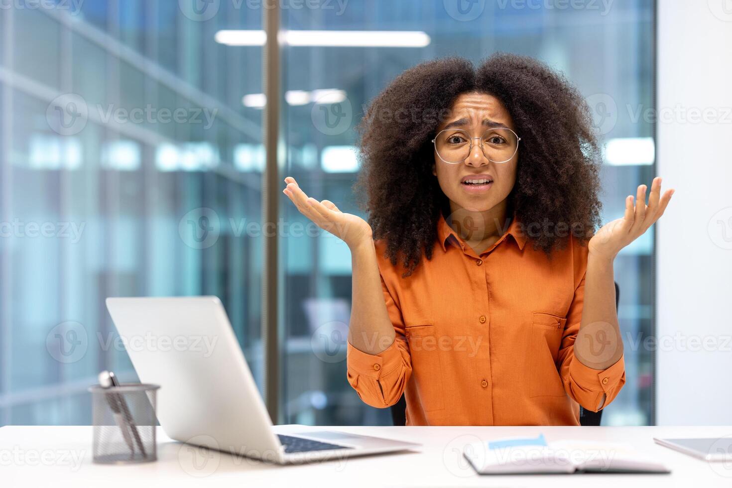 Confused woman in office setting expressing uncertainty. Dressed in orange shirt, sitting at desk with open laptop and book, raising hands in confusion, seeking help. photo