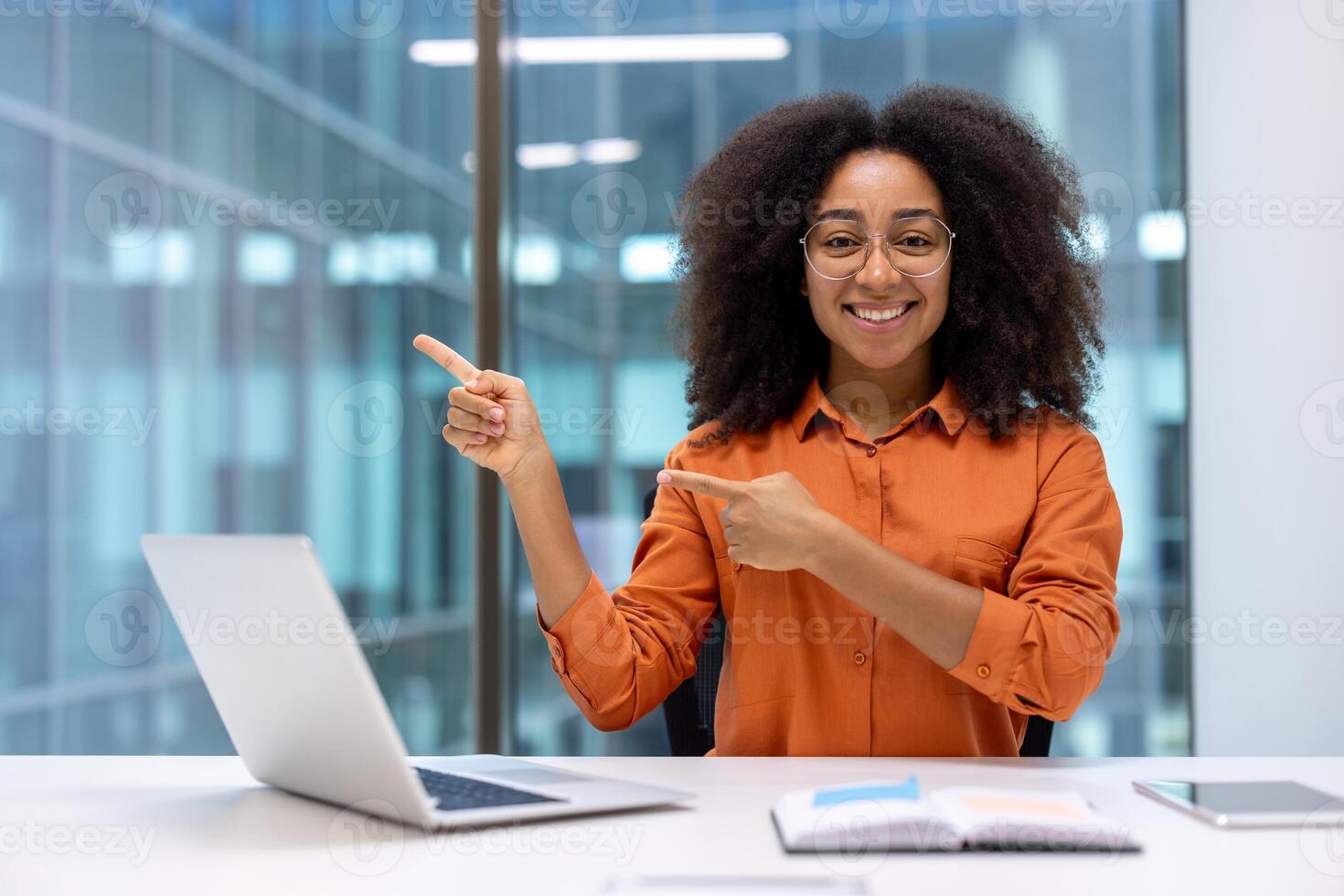 Confident businesswoman sitting at desk in office, pointing at laptop screen with both hands, expressing positivity and engagement. Professional setting, smiling woman, casual business attire. photo