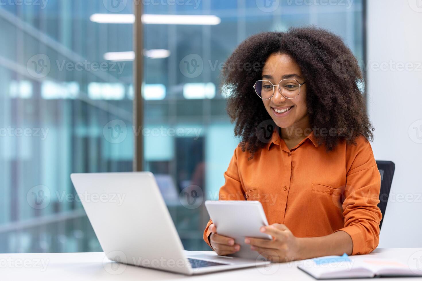 Smiling woman in orange shirt works on digital tablet at desk with laptop. Professional setting in modern office environment with focus on technology and digital connectivity. photo