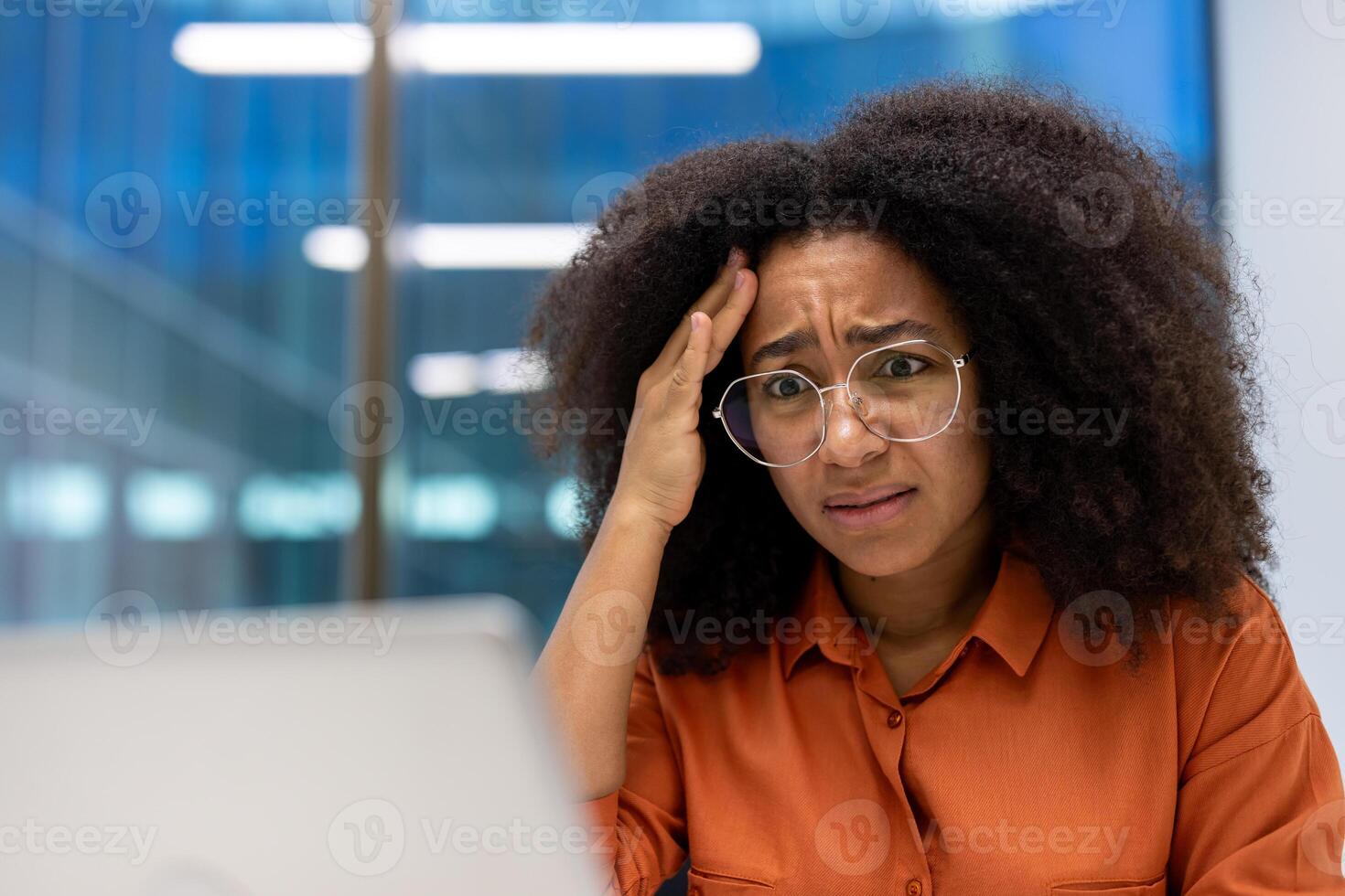 Young African American businesswoman appearing stressed while working at desk in modern office. She wears glasses and orange shirt, expressing concern and worry. Bright setting enhances mood. photo