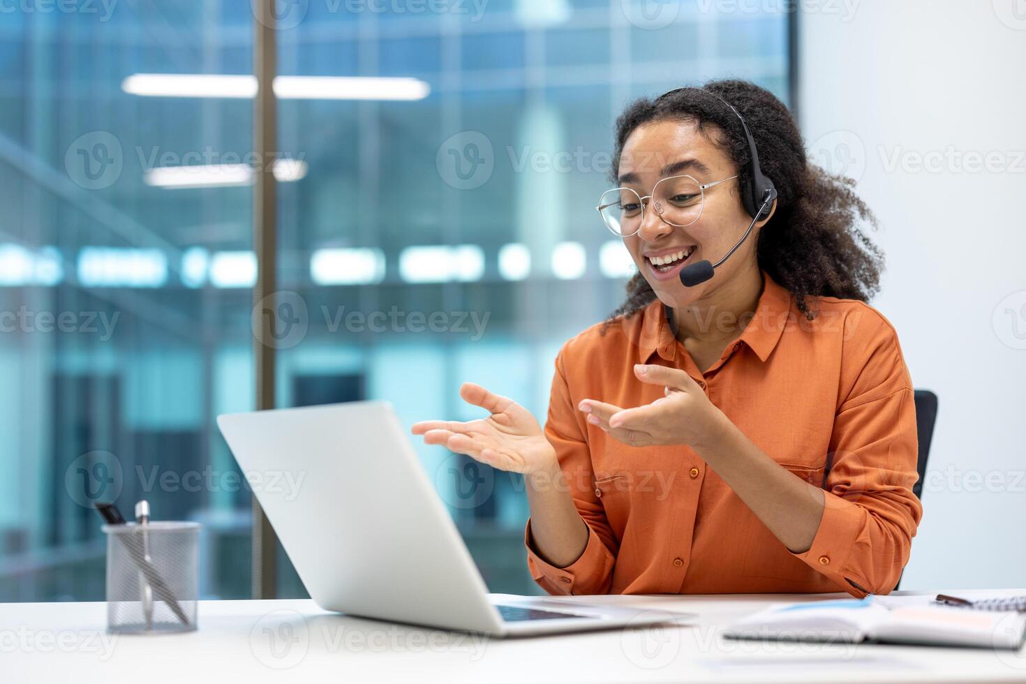 A cheerful businesswoman wearing a headset engages in a conference using her laptop. She gestures expressively while seated in a modern office space, emphasizing connectivity and communication photo