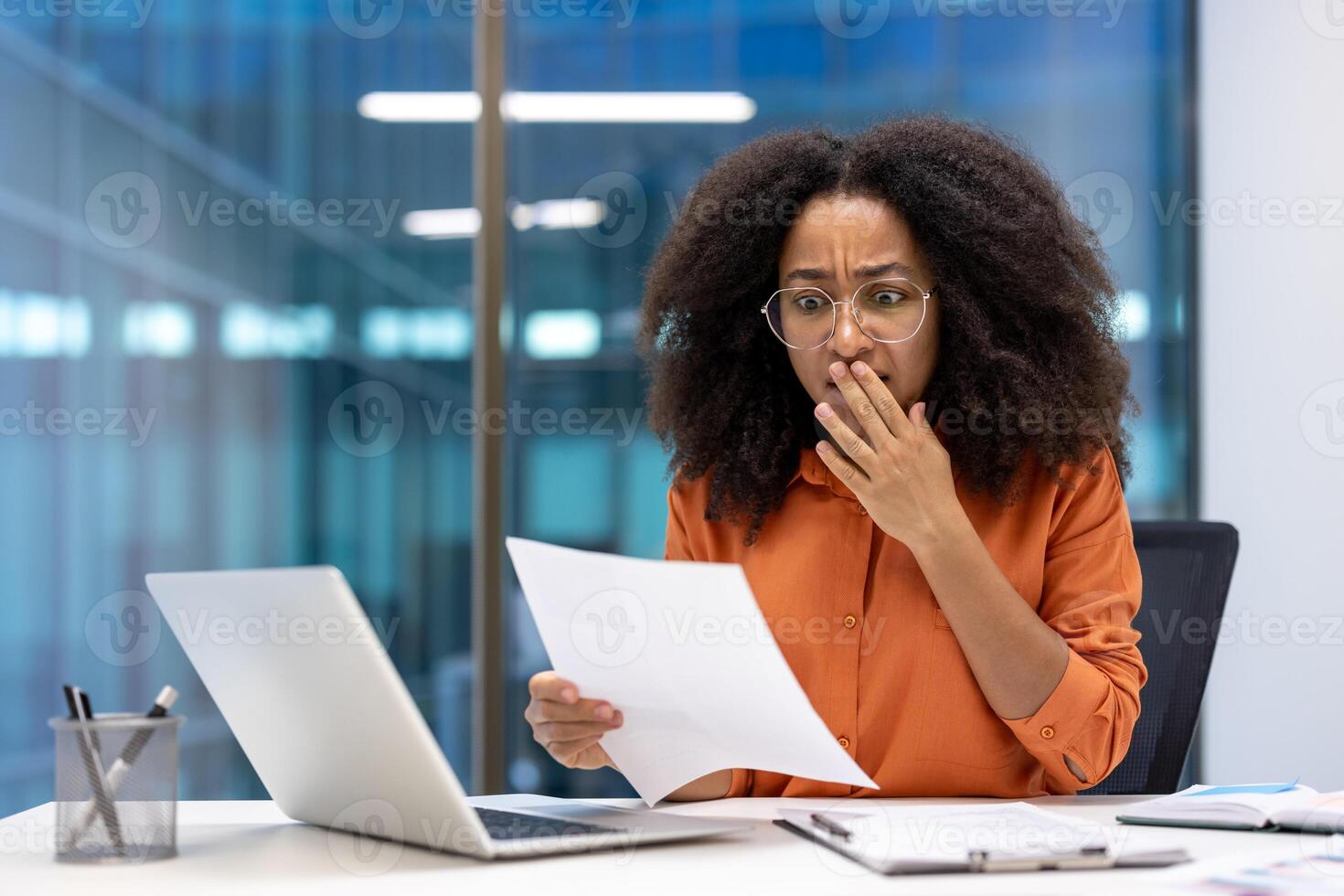 Young African American businesswoman in office expressing surprise while reading document at desk with laptop. Captures emotions of shock and curiosity in professional environment. photo