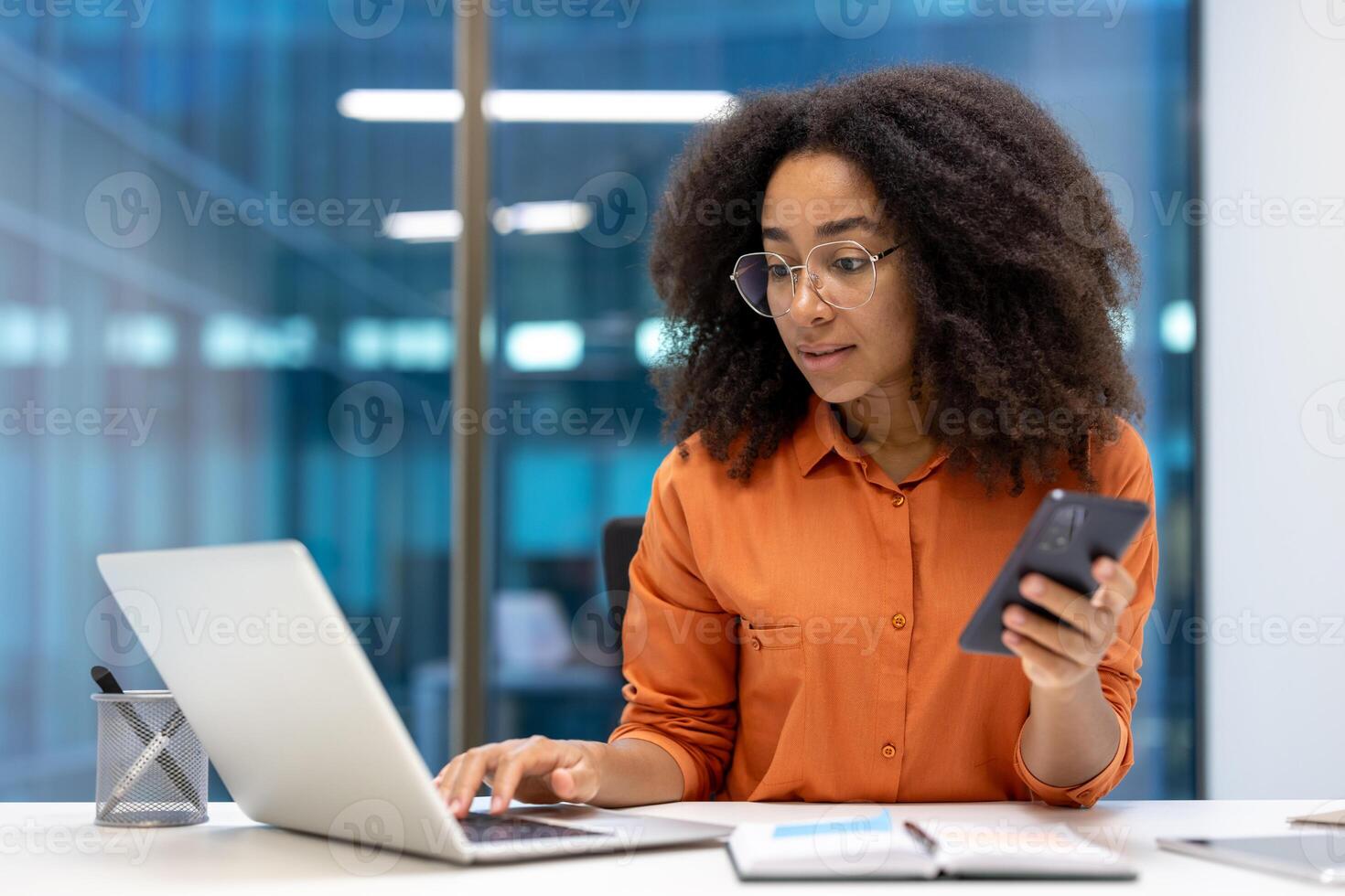 Confident woman working with a laptop and phone in a contemporary office space. She is multitasking by focusing on both devices, showcasing productivity and professionalism. photo