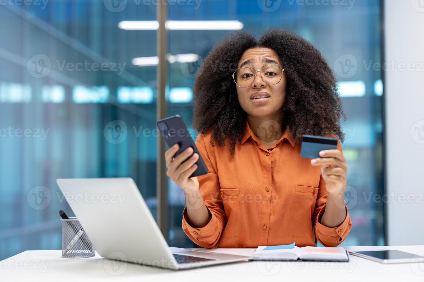 A woman at her desk displaying a smartphone and credit card with a concerned look, suggesting potential financial or technology issues. She is surrounded by a laptop, notepad photo