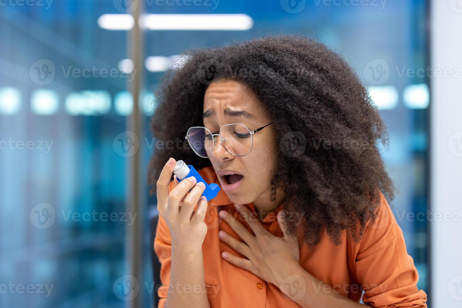 Concerned young woman using asthma inhaler in modern office. She holds her chest, appearing distressed. Office setting visible in background, conveying need for health care support. photo