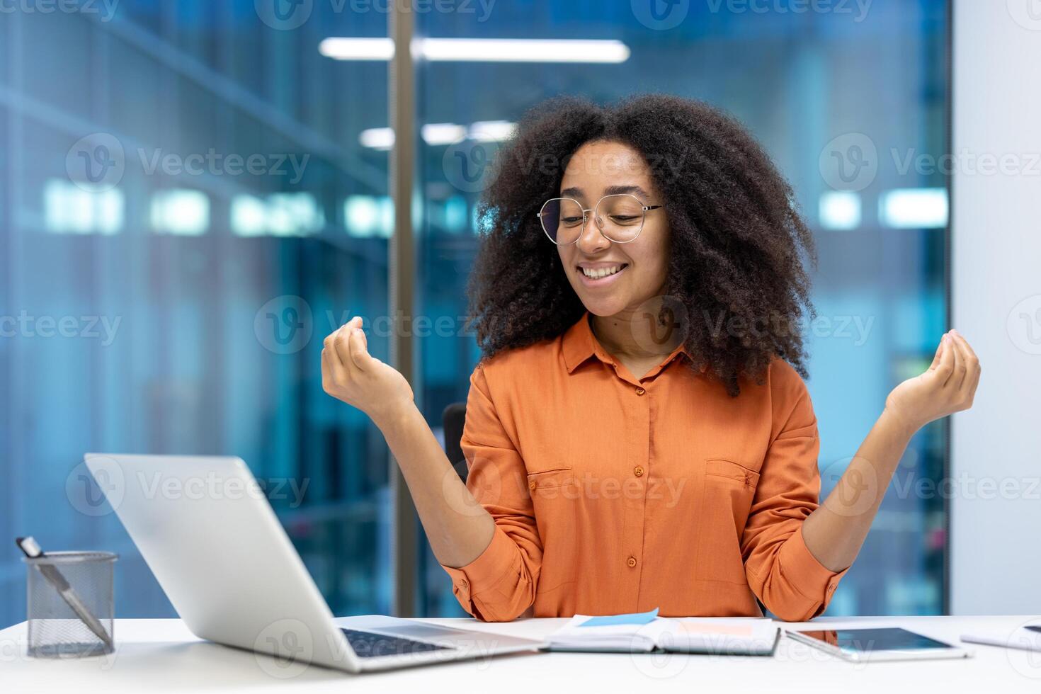 Smiling young woman takes a moment for relaxation and mindfulness during work. She sits at office desk with laptop, phone, and notebook, radiating calm, peace, and balance. photo