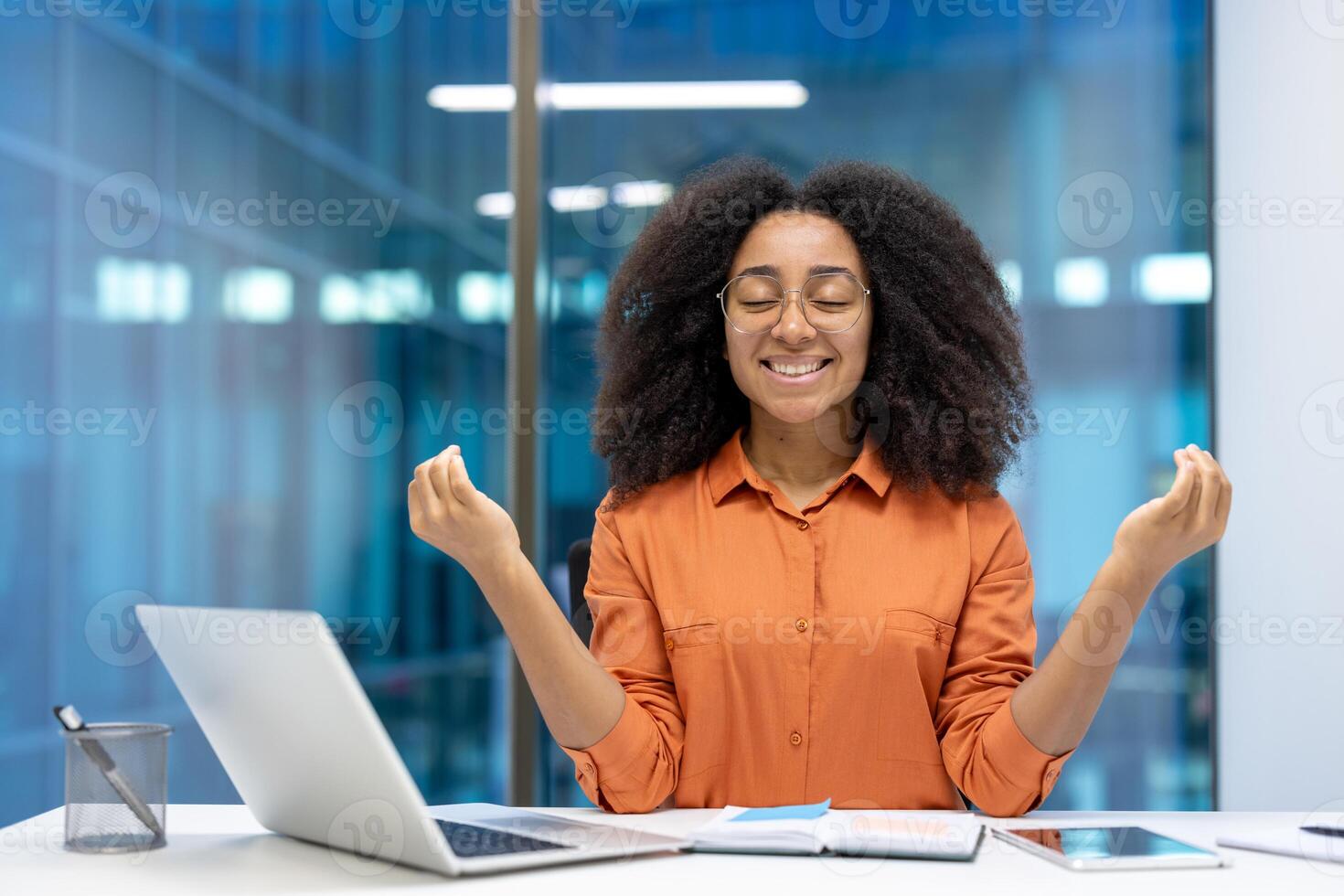 Young African American businesswoman practicing mindfulness in office setting. Relaxed expression, closed eyes, meditative gesture, with laptop and notebook. Concept of stress relief, focus. photo