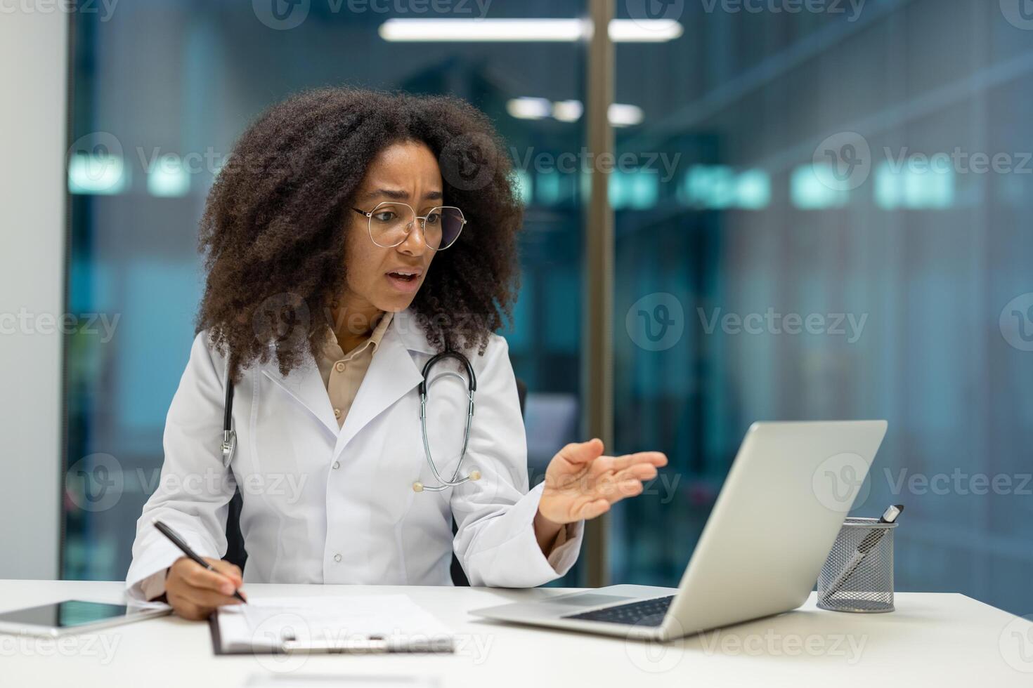 African American woman doctor expressing concern in virtual meeting setting. Seated at desk with laptop, notepad, and pen. Captures telemedicine challenges and professional stress. photo